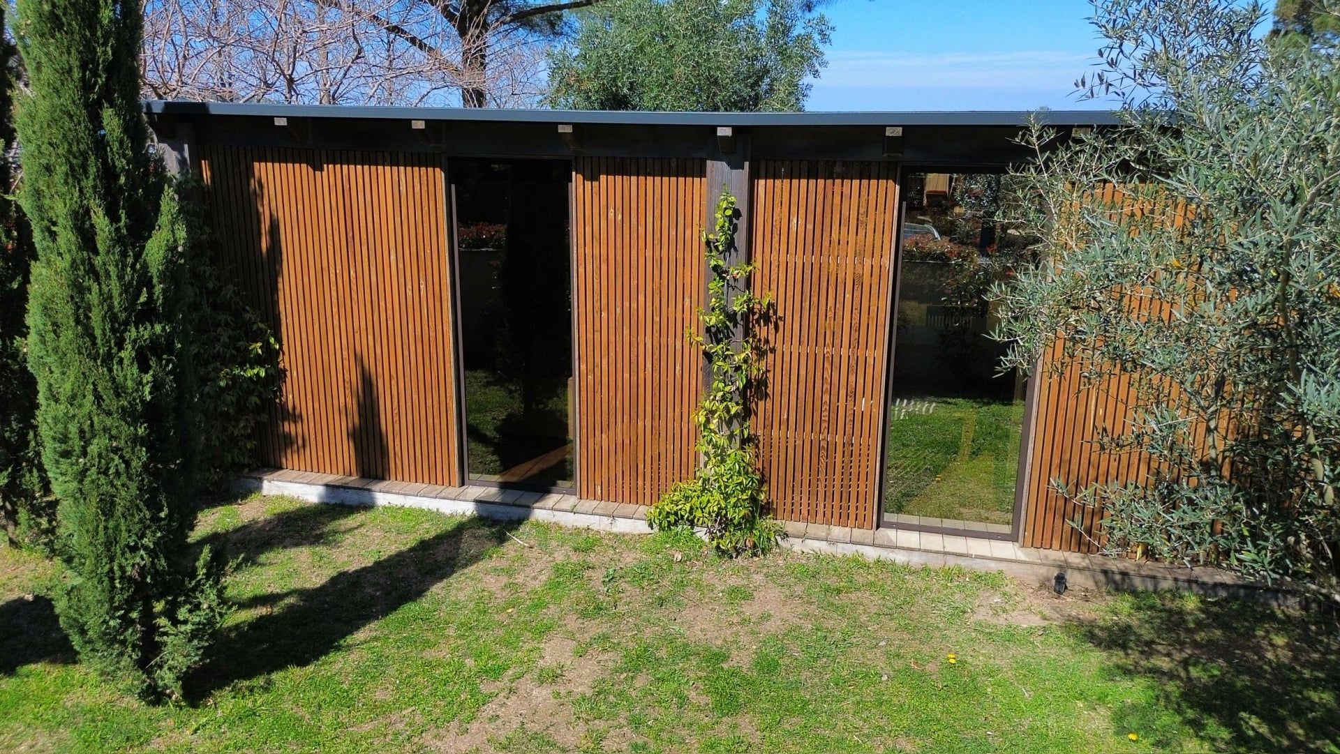 Annexe en bois à bardage vertical et grandes baies vitrées, vue du ciel dans un jardin arboré
