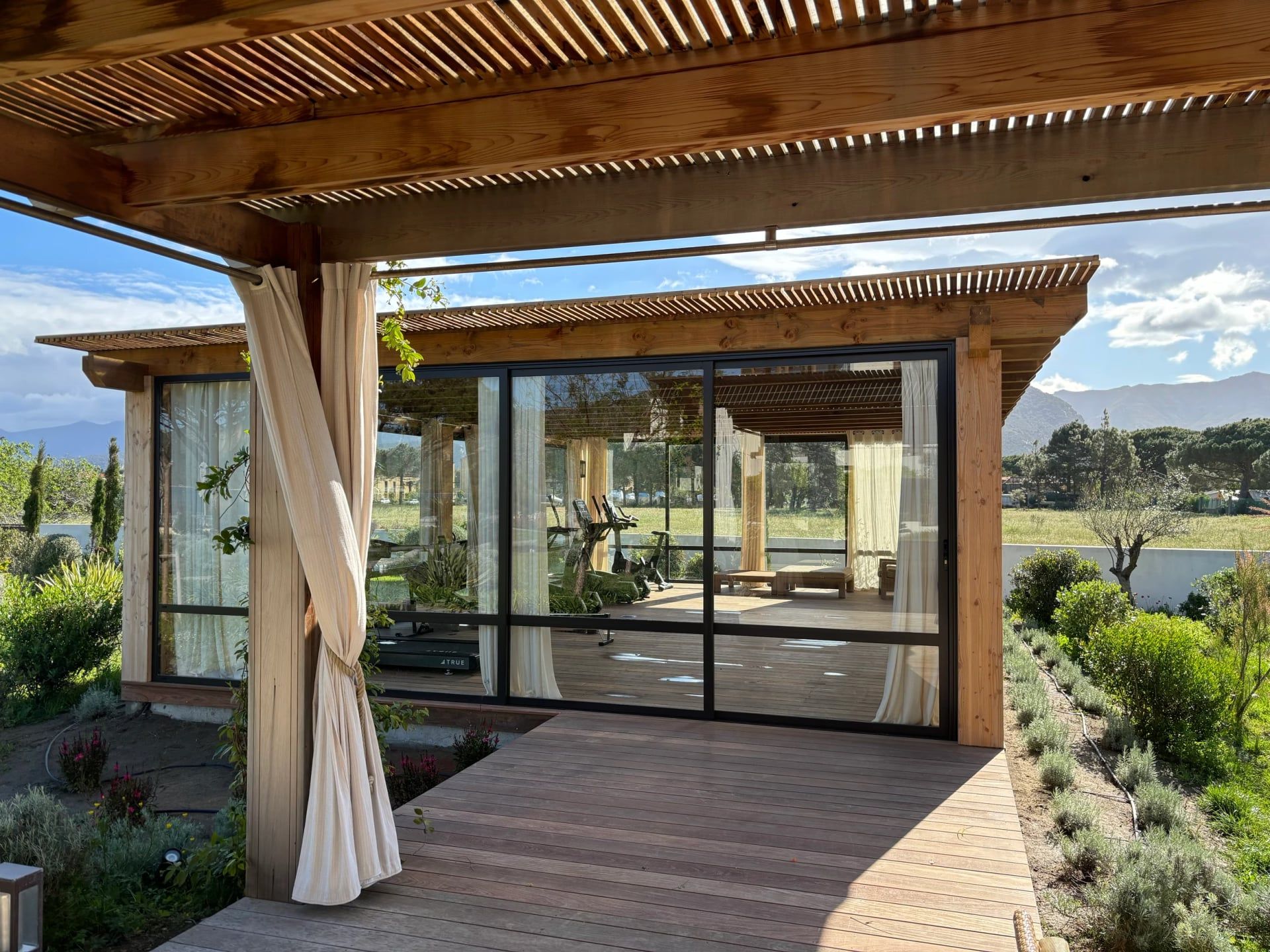 Pergola en bois et terrasse en lames menant à un pavillon vitré, montagnes en arrière-plan